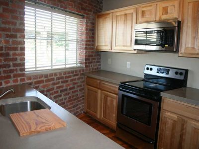A kitchen in Big Lick Junction with hardwood floors, exposed brick walls, stainless steel appliances, and blonde wood cabinetry
